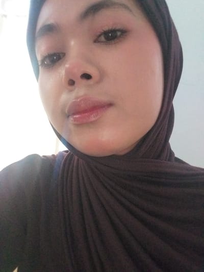 A close-up, head-and-shoulders, selfie-style photograph captures a young woman wearing a dark brown hijab. The subject is positioned centrally in the frame, with her face angled slightly towards the viewer's left. Her eyes are open, and she appears to be looking directly at the camera. She has on light-colored makeup, including eyeshadow and lip gloss, which gives her lips a glossy appearance. The background is out of focus and appears to be a plain, light-colored wall with a bright light source to the left of the frame, suggesting the photograph was taken indoors near a window or light. The image was taken in the city of Watampone, Indonesia.