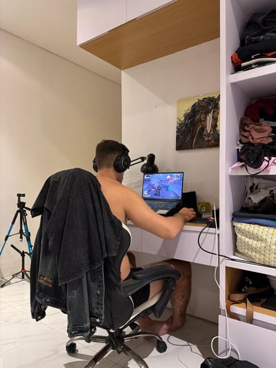 A man wearing headphones sits at a desk playing a video game on a laptop. He is in a room with a white wall and a closet filled with clothes. A painting of a horse hangs on the wall above the desk. A tripod with a camera is visible in the background. The floor is tiled. This scene is located in the city of Kerambitan, Indonesia.