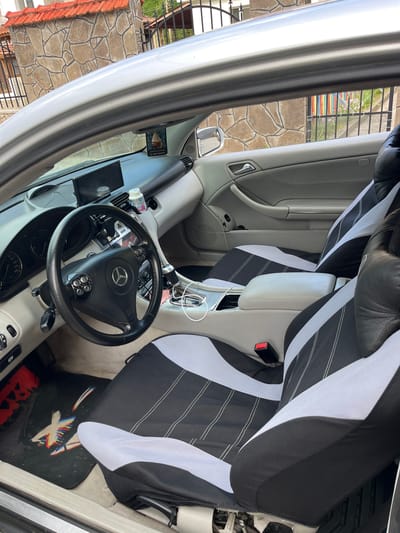The image shows the interior of a silver car, parked outdoors in what appears to be a residential area in Elhovets, Bulgaria. The car is facing forward, with the driver's side door open. The interior is visible, including the dashboard, steering wheel, center console, and front seats. The seats are covered with black and white seat covers. A car air freshener hangs from the rearview mirror. Outside the car, a stone wall and a metal fence are visible. The lighting suggests it is daytime.
