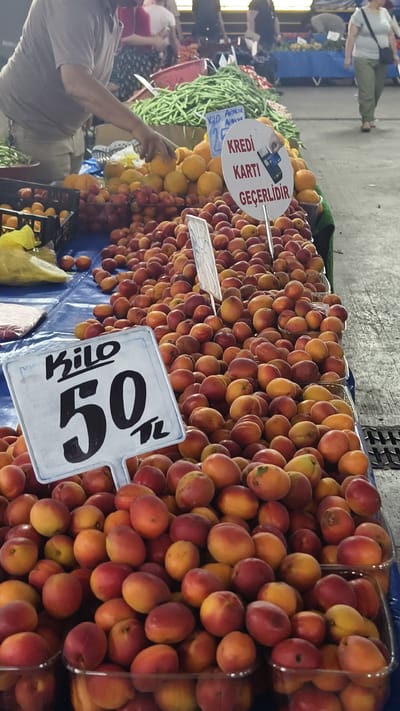 At an outdoor market, likely in Kuşadası, Turkey, a vendor is selling fresh produce. The foreground is filled with a large pile of apricots, with some nectarines or peaches mixed in. A white sign with "Kilo 50 TL" indicates the price per kilogram. In the background, a person is reaching for oranges, and more apricots are visible in plastic containers. Another sign, this one round and white, displays "KREDİ KARTI GEÇERLİDİR," meaning "Credit card is valid." This sign also includes an illustration of a credit card being swiped. Beyond the fruit stands, there are piles of green beans and other vegetables, and a woman wearing a grey top and green pants is walking away from the camera. The lighting suggests it is daytime, and the overall atmosphere appears to be a bustling marketplace.