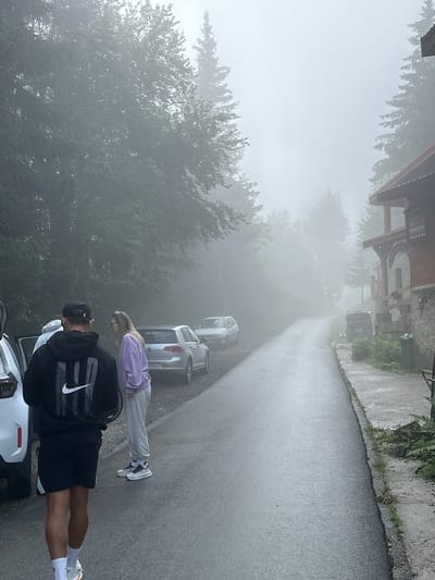 In the city of Trešnjevik, Montenegro, on a foggy day, a group of people stand by a paved road. A man wearing a black hoodie with a white Nike logo and black shorts walks away from the camera. Beside him, a woman in a purple hoodie and white sweatpants stands looking to the left. Behind them, a silver Volkswagen Golf is parked, and further down the road, another white car is visible. A rustic, two-story building with a red-tiled roof and stone facade stands on the right side of the road. The building has a wooden porch with pillars and a sign that reads "WELCOME". The road curves into the distance, disappearing into the dense fog that obscures the surrounding evergreen trees.