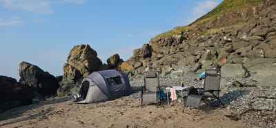 A campsite is set up on a pebble and sand beach at the base of a rocky cliff, with a blue sky overhead. In the foreground, two grey, foldable lounge chairs face a small white folding table. A grey pop-up tent is pitched to the left of the chairs, with its entrance open. On the table, there are a few bottles and a towel. The rocky cliff face is a prominent feature in the background, with sparse vegetation growing on the upper slopes. The beach is strewn with rocks and seaweed. The scene is tranquil and appears to be a location for outdoor recreation. This is likely Girvan, United Kingdom, given the coastal setting and rocky terrain common to the area.