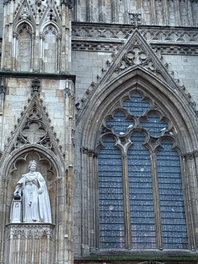 This image captures a portion of an intricately designed building exterior, potentially a cathedral or significant historical structure. The frame is dominated by ornate stonework, including pointed arches, decorative carvings, and a large multi-paned window. A statue of a figure is visible within one of the architectural alcoves. The stonework is weathered, hinting at the age of the building. The presence of birds adds a touch of nature to the scene.