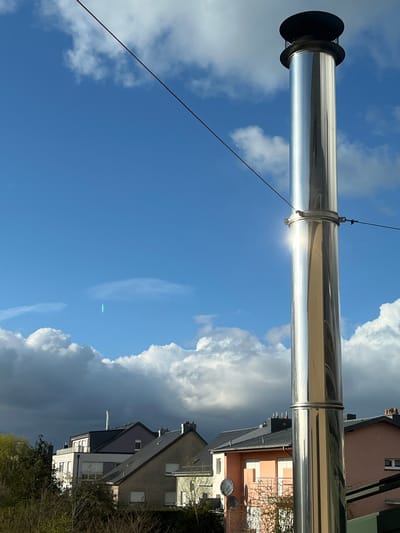 A vertical, polished stainless steel chimney pipe stands prominently on the right side of the frame, reflecting direct sunlight with a bright vertical glare. The chimney is topped with a black cap and features a silver clamping mechanism mid-section, from which a thin black wire extends diagonally across the upper portion of the image. The scene is set in Foetz, Luxembourg, under a partially cloudy sky, which displays areas of bright blue interspersed with white cumulus clouds and darker grey clouds at a lower altitude. Below the sky, the rooftops and upper sections of multiple residential or multi-story buildings are visible. These include a building with a dark grey roof and white facade, and another with a light pink facade and dark roof, with two black satellite dishes mounted on a pole adjacent to it. Smaller, white chimneys are discernible on distant rooftops. Sparse tree canopy is present in the lower-left corner. A faint, vertical turquoise-green light streak is visible in the clear blue sky in the upper-left quadrant. No individuals or dynamic actions are depicted.