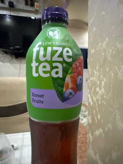 A close-up, eye-level shot shows a green plastic bottle of Fuze Tea Forest Fruits with a purple cap. The bottle is partially filled with a dark amber liquid. The label features a white logo with "fuze" in large letters, "tea" below it, and "BLACK TEA EXTRACT" and "LOW CALORIES" text. A graphic of mixed forest fruits, including blueberries and raspberries, is visible on the label. The bottle rests on a white surface, possibly a table, covered with a checkered tablecloth. In the background, a blurred television screen is visible to the left, and a textured, cream-colored wall or curtain is on the right. The scene appears to be indoors, possibly in a home environment. There are no people visible in the frame. The location of Samuilovo, Bulgaria, is not discernible from the visual information provided in the image.