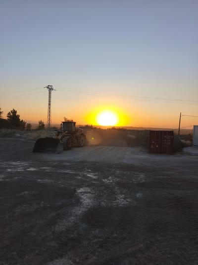 The image depicts a construction site in Riba-roja de Túria, Spain, at sunset. A large yellow front-end loader is positioned on a gravelly surface, its bucket lowered. In the background, a red shipping container sits near a pile of dirt and gravel. The sun, a bright, intense yellow orb, is low on the horizon, casting a warm orange and yellow glow across the sky and landscape. Electrical pylons and wires are visible against the fading blue and orange sky. The ground in the foreground appears to be wet in patches, reflecting the light. Sparse vegetation and trees dot the middle ground, with distant hills silhouetted against the horizon. The scene has a raw, industrial feel, illuminated by the dramatic natural light of sunset.