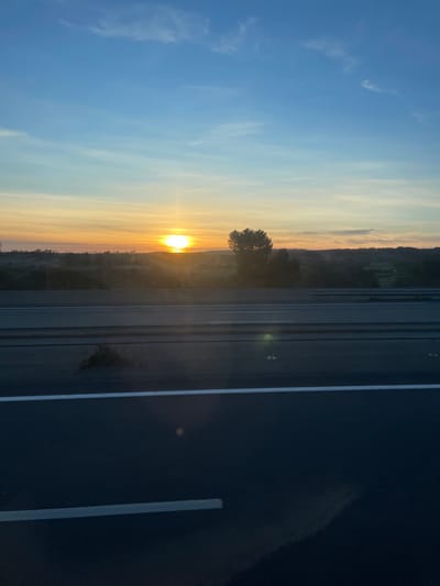 The image depicts a sunset observed from a vantage point overlooking a multi-lane highway in Sigean, France. The sky, occupying the upper two-thirds, presents a gradient from pale blue at the zenith to vibrant orange and yellow near the horizon, indicating the setting sun. Sparse cirrus clouds are visible against the blue sky. The sun, a brilliant disc, is positioned slightly left of the image center along the horizon, emitting a strong lens flare and illuminating the atmosphere with warm hues.

Below the horizon, the distant landscape consists of rolling hills and dense tree lines, appearing as dark silhouettes against the bright sky. The foreground, occupying the lower third of the image, features a dark asphalt highway with distinct white lane markings. A concrete or metal barrier/guardrail runs horizontally across the middle ground, separating the highway from the distant landscape. At least two lanes of traffic are visible on the highway. No individuals or vehicles are identifiable within the scene. Light reflections are present on the road surface.