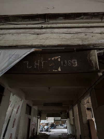 This image depicts a deep, narrow, and neglected vertical shaft or passage within an old building in Bratsigovo, Bulgaria. The rough, concrete walls are heavily discolored and textured, showing signs of peeling paint, grime, and overall deterioration. Exposed pipes, electrical conduits, and wires crisscross the surfaces, emphasizing the building's age and state of disrepair.
A prominent feature on one side is a dark, rusty metal door or panel. Faded white markings are visible on its surface: the number "6301" is clearly identifiable at the top, and below it, the letters "LH2" (or "L H Z") are written vertically.
The ceiling consists of exposed concrete beams and appears unfinished or damaged, with some debris present. Looking further down into the shaft, the floor below reveals several open rectangular hatches or access points. A significant portion of this floor is covered by a white, translucent plastic sheeting. This sheeting bears a repeating "W M" logo, the web address "www.WMHols.com", and possibly the word "DELEEP," indicating it is a protective packaging material.
At the very bottom of the passage, a cluttered area is visible, filled with stacked cardboard boxes, what appears to be a dismantled wheelchair, and other miscellaneous items, suggesting a storage or utility space. The lighting is dim and uneven, creating deep shadows that enhance the gritty and somber atmosphere. No people are present, and the scene conveys a strong sense of decay and abandonment within an old, possibly industrial or utilitarian structure.