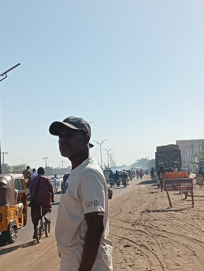 A man wearing a baseball cap and a QNB t-shirt stands on a dusty roadside. In the background, a busy street scene unfolds with motorcycles, auto-rickshaws, and other vehicles moving in both directions. A man rides a small bicycle beside an auto-rickshaw. A sign for 'GARBA mai battery' is visible near a truck. Utility poles line the road under a clear, bright sky.