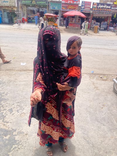 The image shows a young boy being carried by a woman down a street in Kuchlak, Pakistan. The boy wears a purple cap, a black top with an orange sleeve detail, and looks directly at the camera. The woman is draped in a black shawl with pink embellishments and a patterned maroon and beige dress. They are on a paved street with multiple shops lining the background, including food stalls with signs in Arabic script and names like "Sweet Corner" and "BisMillah Hotel". Pedestrians and parked vehicles are also visible in the background.
