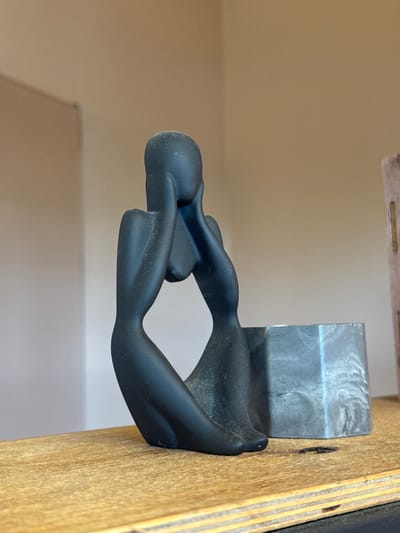 This is a close-up, vertically oriented, eye-level shot of a black, abstract sculpture resembling a human figure in a seated or reclining pose. The sculpture is made of a matte, dark material. It is attached to a light-colored wooden surface, which has visible grain and some imperfections. To the left of the sculpture, a metallic object with a brushed or textured surface is also visible, seemingly part of the mounting or display. The background is a plain, neutral-colored wall, out of focus. The lighting is soft and appears to be from an indoor source. There is no visible text in the image. The scene appears to be a still life or an indoor display, possibly in a studio or a home setting. The orientation of the image suggests it might have been taken with a phone camera.