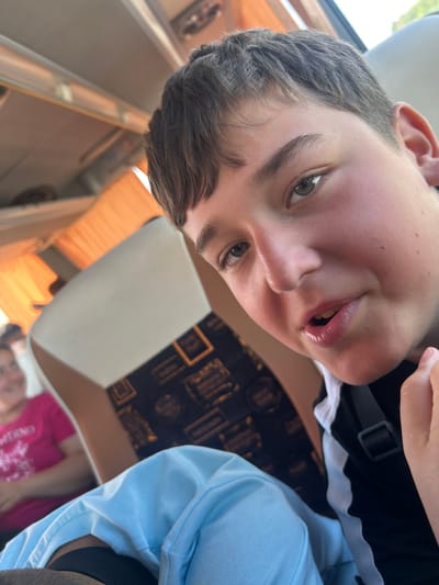A close-up, selfie-style shot captures a young boy with short brown hair and light green eyes looking at the camera. He appears to be in a vehicle, possibly a bus, with seats visible in the background. The boy is wearing a black shirt with white trim and a grey strap. A light blue garment is in the foreground. To the left and out of focus, a woman in a pink shirt is seated. The overall environment suggests an indoor setting with artificial lighting. The location is Polsko Padarevo, Bulgaria.