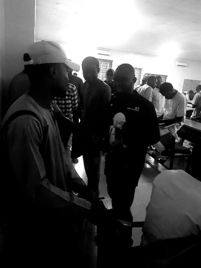 This black and white image, captured in Panhauya, Nigeria, depicts an indoor gathering of approximately ten to fifteen male individuals, likely within a classroom or meeting hall. In the immediate left foreground, a man wearing a light-colored baseball cap and a long-sleeved, light-toned garment stands, facing towards the right with his profile visible. Adjacent to him, in the central foreground, another man wears a dark, short-sleeved shirt with the letters "R/F" and additional text visible on the left chest. This individual is holding a small, light-colored doll or figurine against his torso. The interaction between these two foreground figures is not explicitly clear.
In the midground and background, multiple other individuals are present. Some are standing, while others are seated at desks or tables on the right side of the frame, suggesting an organized setting. Several background figures wear light-colored traditional attire, and one individual in the midground wears a plaid or checkered shirt. The environment includes light-colored walls, a bright ceiling with visible light fixtures, and an air conditioning unit mounted on a far wall. A patterned or textured surface, possibly window blinds or wall covering, is visible in the mid-background. The scene is characterized by significant shadow in the foreground and lower portions, indicating overhead or directional lighting.