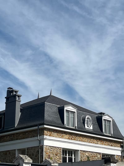 The image shows the roof and upper facade of a stone building under a cloudy blue sky. A dark grey slate mansard roof with two peaked dormer windows and one circular dormer window is prominent. A tall, grey, stepped chimney is visible on the left side of the roof. The building's facade is constructed of rough, irregularly shaped stones in shades of brown and orange, bordered by a white decorative band. Below this, more windows are partially visible. The scene appears to be an exterior view of residential architecture. Based on available information, this building is located in Chevilly-Larue, France.