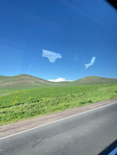 The image depicts a scenic landscape viewed from what appears to be inside a vehicle, possibly a bus or car, given the reflections on a surface, likely a window. The scene is dominated by a vast expanse of clear blue sky, interrupted by a few wispy white clouds.

On the right side of the frame, a sloping, lush green hill covered in grass and scattered yellow wildflowers rises. Adjacent to the hill is a paved road with a white line marking its edge. The road surface is grey and appears to be asphalt. The perspective suggests the vehicle is moving alongside the hill and the road.

Reflected on the surface, likely a window, are abstract shapes of light and possibly interior elements of the vehicle, including what appears to be a white, geometric object. The overall lighting indicates it is daytime, with bright sunlight. The vibrant green of the vegetation and the clear blue sky suggest pleasant weather conditions. The absence of people or distinct man-made structures in the wider landscape, beyond the road, points to a more rural or natural setting, possibly in a mountainous or hilly region. The rotation of the image suggests it was taken with the camera held in a portrait orientation, and the content is presented sideways. No visible text is present in the image.