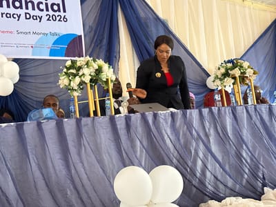 An indoor event, possibly a conference or seminar, is captured with a woman speaking at a podium or head table. The woman, dressed in a black suit with a red top and a decorative brooch, stands centrally, gesturing with her right hand as she addresses an unseen audience. A MacBook and a microphone are on the table in front of her.

Behind the table, several individuals are partially visible, including a man with a beard holding a microphone and another man wearing glasses. Two elaborate floral arrangements featuring white and blue flowers on gold stands adorn the table, alongside multiple water bottles (one clearly labeled "SWAN") and a small white fan.

The setting is decorated with white and blue draped fabrics in the background, suggesting a formal event space. A large banner hangs behind the speakers, prominently displaying "Financial Literacy Day 2026" and the "Theme: Smart Money Talks." Below the theme, contact information and social media handles for "@NDICNigeria" are visible, providing a strong location cue to Nigeria, specifically Unguwan Dosa. White balloons are scattered throughout the foreground and background, adding to the celebratory or event atmosphere. The overall impression is a professional and engaging public speaking event focused on financial education.