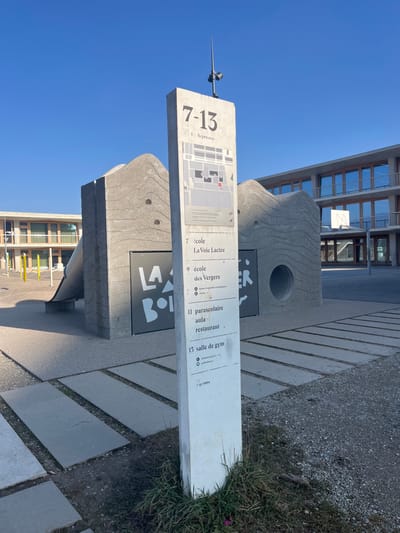 An outdoor scene captures a large concrete information sign and an adjacent sculpted stone structure under a clear blue sky. No people are visible.

The main sign is a tall, slender, light grey concrete monolith oriented vertically. On its upper left, it displays the large numbers "7-13" and "1. Arpenaz" below them, accompanied by a map diagram. The right side of the sign lists destinations and their corresponding numbers: "7 école La Voie Lactée," "9 école des Vergers," "11 parascolaire aula restaurant," and "15 salle de gym." Near the bottom of this list, text reading "RETEIN" and "MEYRIN" is visible. A small pointed antenna-like object is affixed to the top of the sign.

Next to this sign is a substantial, darker grey concrete structure with a wavy, organic design and several circular cutouts, one of which is prominent. Dark square panels are set into this structure, displaying partial text. "LA" is visible on one panel, and "BOI" is legible on another.

In the background, a modern building with large glass windows and a visible basketball hoop suggests a school or public complex. The ground is a mix of patterned grey pavers and sparse grass. The clear sky and bright light indicate it is daytime with sunny weather. The location context indicates this is in Meyrin, Switzerland.