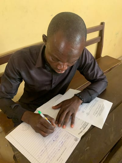 A male individual with short dark hair and a beard is seated at a wooden desk, actively writing on a lined paper with a green transparent ballpoint pen held in his right hand. He is wearing a dark, long-sleeved collared shirt and a dark beaded bracelet on his right wrist. His fingernails are painted a light reddish-orange hue. Adjacent to the lined paper is a document partially titled "LYTECHNIC LOKOJA APPLICATION FORM." The background features a plain light yellow wall and a section of a brown wooden chair or bench. The scene is indoors. The location is contextualized as the city of Abobo, Nigeria, with a visible document referencing "LYTECHNIC LOKOJA."