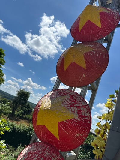 Three conical hats decorated with red and yellow stars, likely representing the Vietnamese flag, are mounted vertically on a metal frame against a bright blue sky filled with scattered white clouds. The hats are positioned at varying distances from the camera, with the closest one in the foreground and the farthest in the upper right corner. The hats appear to be made of woven material and show signs of wear, with chipped paint and weathered textures. In the background, a lush green landscape with trees and rolling hills is visible, suggesting a rural or semi-rural setting. To the right of the hats, a cluster of artificial yellow flowers adds a decorative element. The scene is illuminated by bright daylight, casting subtle shadows and highlighting the textures of the hats and surrounding foliage. The location is specified as Ia Hru, Vietnam.