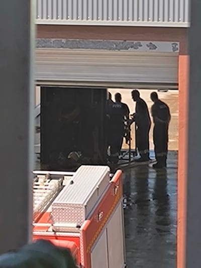 The image captures a scene within what appears to be an emergency services facility, likely a fire station, in Vilafranca del Penedès, Spain. Three male figures, silhouetted against a bright background, are gathered inside a large bay or garage opening. They are dressed in dark uniforms, possibly firefighter turnout gear or work coveralls, with one figure on the left showing a faint, light-colored patch or emblem on his back. They appear to be interacting with or inspecting a piece of equipment, possibly a portable stand or tool, positioned between the two central figures.
In the foreground, partially visible on the left, is the top section of a red and white emergency vehicle, consistent with a fire truck. It features a checkered pattern on a storage box or compartment. The floor of the bay is dark and highly reflective, suggesting it is wet or freshly cleaned.
Above the large opening, a multi-layered wall construction is visible. It includes a section of corrugated metal siding at the very top, followed by a brown horizontal band, a textured grey band, and then a white horizontal strip directly above the garage door. On this white strip, there are faint, partially obscured, and possibly faded or damaged dark markings. While indistinct, these markings appear to be text or a logo. On the left side of the strip, some fragmented characters are visible, potentially part of a word like "GOMAR" or similar. A smaller, blurry graphic or logo is present on the right side of the strip.
The bright lighting suggests it is daytime, casting the figures into silhouette. No specific emotions are discernible. The overall setting implies a period of readiness or maintenance within an emergency response context.