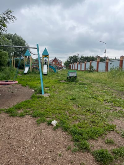 This image depicts a children's playground located in the city of Nagayevo, Russia. The playground features a blue and yellow multi-level play structure with two slides, a climbing area, and a swing set with a circular tire swing. In the background, there are several residential buildings with red roofs and a long fence made of metal and brick. The ground of the playground is a mix of dirt and overgrown grass. A small green trash receptacle is visible near the fence. The sky is overcast with gray clouds.