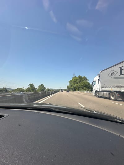 This image captures a view from the interior of a vehicle traveling on a highway. The perspective is from the driver's seat, looking forward and slightly to the right. The foreground is dominated by the dashboard of the car, a dark, textured surface. Beyond the dashboard, the highway stretches into the distance under a clear, bright blue sky.

On the right side of the highway, a large white truck with a grey trailer is visible. The trailer has a prominent black logo featuring the letters "IB" enclosed in a diamond shape. The truck appears to be in motion, traveling in the same direction as the car. Other vehicles, including cars, are also present on the highway.

To the left of the highway, there is a concrete barrier, and beyond that, a landscape with trees and rolling hills. Some of the trees are lush green, suggesting a temperate climate and likely springtime or summer. In the distance, there are indications of a town or city, with structures and infrastructure visible on the horizon. There is a plume of dark smoke rising from behind a cluster of trees on the left side of the highway, suggesting a fire or industrial activity.

The sun is high in the sky, casting bright light and creating some glare and reflections on the windshield, as evidenced by the bright spots in the upper portion of the image. The overall impression is of a sunny day on a highway journey.

While the exact location within Mercurol-Veaunes, France, cannot be determined from the image alone, the scene depicts a typical highway environment in a rural or semi-rural area, consistent with regions in France. The presence of the highway, surrounding vegetation, and distant structures are common features of such locations.