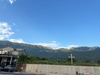 The image depicts a scene in Radanovići, Montenegro. In the foreground, a concrete barrier runs horizontally across the frame, with some graffiti visible on its surface. To the left of the barrier, a dark-colored station wagon is parked next to a building with an open facade and large windows reflecting the sky. Behind the building, a steep, green hillside rises. In the midground, a white cross stands amidst overgrown vegetation. The background is dominated by a range of lush, green mountains under a clear blue sky with scattered white clouds. A faint contrail from an aircraft is visible in the upper portion of the sky.
