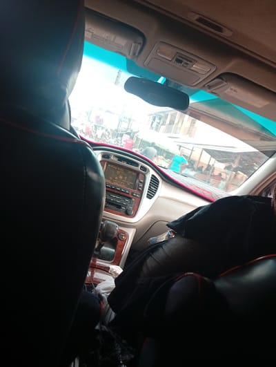 The image is taken from the interior of a car, looking forward through the windshield at an outdoor scene in the city of Ukpor, Nigeria. The foreground is dominated by the dark interior of the car, with the back of a black leather seat with red trim visible on the left and a portion of another seat on the right. The car's dashboard is visible, featuring a digital display and radio controls. Outside the car, through the windshield, a busy street scene unfolds. People are visible, some walking and some gathered around stalls or what appear to be market activities. Buildings, possibly shops or houses, line the street. The overall impression is one of a bustling urban environment. The lighting suggests it is daytime, with bright sunlight casting shadows.
