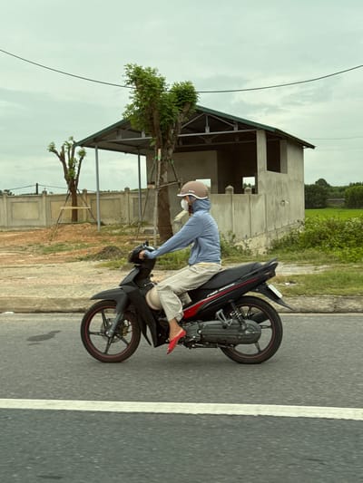A person wearing a blue hoodie, a light-colored face mask, and a light brown helmet rides a black motorcycle on an asphalt road in Ái Tử Commune, Vietnam. The rider is wearing striped pants and red shoes. Behind the motorcycle, there is a partially constructed building with a green metal roof, a concrete fence, some dirt piles, and sparse vegetation. In the background, there are trees and a field of green crops under a cloudy sky. A power line stretches across the upper portion of the frame.