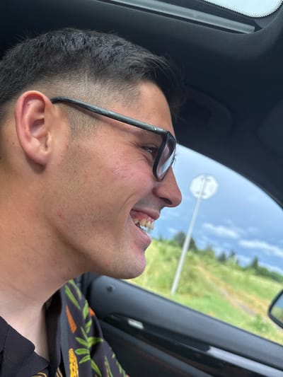 A man is captured in a close-up, profile view from inside a vehicle, looking upwards and to the right. He appears to be laughing or cheering, with his mouth open, revealing his upper teeth. He wears dark-rimmed glasses and a black shirt adorned with a distinctive pattern of green leaves and orange, leopard-like spots. His hair is dark and neatly cut on the sides.
The setting is inside a car, with elements of the dark interior, such as the headliner and window frame, visible. Through the car window, a bright, clear blue sky with scattered white clouds is prominent. A blurred white octagonal road sign on a pole is visible against the sky, suggesting a stop sign. Beyond the sign, a vibrant green, grassy landscape with distant trees can be seen, indicative of a rural or roadside environment. The scene takes place during a sunny daytime, suggesting pleasant weather. The man's expression conveys strong emotion, likely joy or amusement. There is no discernible text visible in the image. The location context for this scene is Svoboda, Bulgaria.