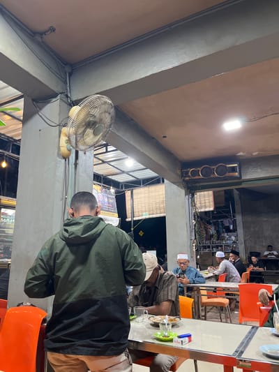 The image depicts an outdoor dining area in Kutalhoksukon, Indonesia. People are seated at tables, some wearing traditional Muslim headwear. One person is wearing a green and black jacket. A fan is mounted on a support beam. A speaker is mounted on the wall. The scene suggests a casual evening gathering, possibly a meal.