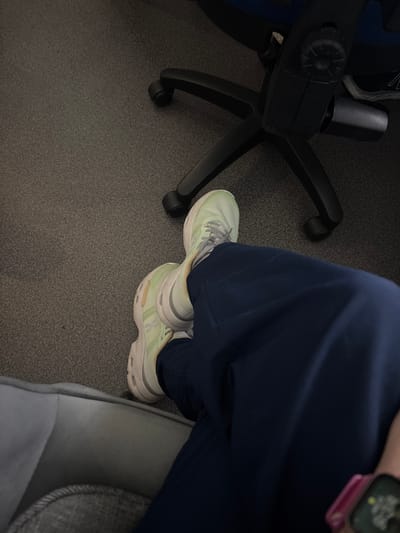 The image shows a partial view of a person seated indoors, with the setting context provided as Medellín, Colombia. The person's lower legs are visible, crossed at the ankles, wearing dark blue trousers or scrubs. On their feet are light green athletic shoes, one resting atop the other. The design of the shoes, particularly the sole with its distinctive pods, suggests they are from the "On" brand, possibly a "Cloud" model. To the left, the black base and leg of an office-style chair with wheels are clearly seen on the gray carpeted floor. To the right, a gray upholstered surface, possibly an armrest or sofa, is present. A digital smartwatch with a pink band is visible on the person's wrist. The scene conveys a moment of rest or waiting in an indoor environment. No specific time of day, weather, or discernible emotions are indicated. There is no clearly readable text apart from potential branding on the shoes.