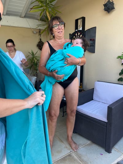 In an outdoor patio setting, likely in Saint-Galmier, France, a woman with glasses and a black swimsuit holds a baby wrapped in a turquoise blanket. The baby has curly dark hair and is looking to the side. Another woman, also wearing glasses and a white t-shirt, stands in the background to the left, gesturing with her hands. To the right, a dark wicker chair with white cushions is visible, and a dartboard hangs on the wall. A third person, whose hands are visible in the foreground, is holding up a large piece of turquoise fabric. The environment suggests a warm, possibly sunny day, with tiled flooring and some potted plants.