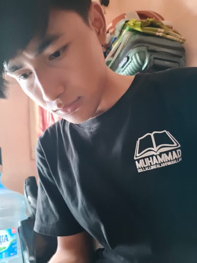 A young man is positioned in an indoor setting, seemingly in Cianjur, Indonesia, based on the "AQUA" water bottle visible in the lower left. He is looking down, wearing a dark t-shirt imprinted with "MUHAMMAD SOLLALLOHU'ALAIHIWASALLAM" and a graphic resembling an open book. The background features a pale peach-colored wall, a stack of items that could be luggage or storage containers, and a partial view of what might be a red curtain or cloth. The man's facial expression is contemplative.