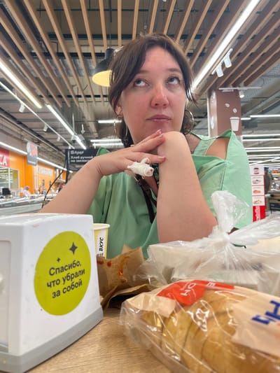 The image shows a woman sitting at a table in what appears to be a cafeteria or food court within a supermarket. She is looking upwards, with her arms crossed and resting on the table. Her right hand has a white bandage wrapped around it. In the foreground, there is a white napkin dispenser with a yellow sticker that reads "Спасибо, что убрали за собой" (Thank you for cleaning up after yourself) in Russian. There is also a paper cup with Russian text, a brown paper bag, and a plastic bag containing sliced bread. In the background, supermarket aisles and signage are visible, including a sign that says "КУЛИНАРИЯ" (Culinary) and another with Russian text about bread. The lighting suggests an indoor commercial environment. The location is identified as Volzhsky, Russia.