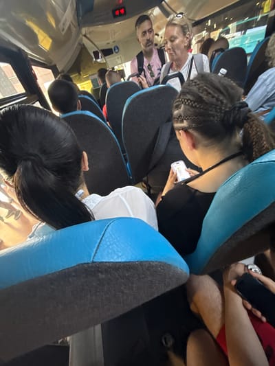 This image depicts the interior of a public transit vehicle, likely a bus or shuttle, in Las Palmas De Gran Canaria, Spain. The perspective is from a rear seat, looking forward and slightly to the right, showing multiple passengers.
In the immediate foreground on the left, the back of a person's head with long, dark, straight hair tied in a high ponytail is visible. This individual wears a white collared shirt. To the right, slightly further back, the shoulders and back of another person's head are seen. This individual has dark hair styled with multiple tight braids on the scalp leading into a single, voluminous, crimped ponytail. They are wearing a dark, possibly black, off-shoulder top and are intently focused on a white smartphone held in both hands, displaying a light-colored screen with application content.
In the midground, on the left side, a man with short, dark hair, a beard, and a mustache stands or leans. He wears a light pink short-sleeved collared shirt with sunglasses hanging from the collar and a dark backpack. He looks towards the front-right. Adjacent to him, a blonde woman stands or leans, wearing sunglasses on top of her head, a white t-shirt, and a grey backpack. Her expression is neutral.
Further into the vehicle, several other passengers are visible from behind, seated in various rows with their backs to the viewer. A small white plastic water bottle rests on top of a seat in the mid-right. In the bottom right corner, the hairy legs of a person wearing red shorts are visible, holding a black smartphone horizontally.
The vehicle's interior features blue and dark grey upholstered seats. Overhead, a red digital display shows the numbers "10 45". The ceiling and overhead panels are a light, possibly white or cream, color. Windows on the left side offer a blurred view of external buildings with reddish-brown and beige tones. A blue circular sign with a white arrow is affixed to an overhead panel on the right side of the vehicle. The floor appears dark. The scene suggests a routine travel environment.