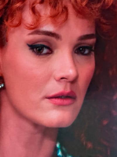 This is a close-up, headshot of a woman with bright red, curly hair. She has fair skin with a pinkish tone, dark eyeliner creating a winged effect, and pink lipstick. A small, shimmering earring is visible on her left ear. Her gaze is directed slightly to the left of the camera. The background is dark and out of focus, with hints of her curly hair extending to the right. The image appears to be a still from a film or a portrait, with a soft, warm lighting that emphasizes the subject's features. The specific location in Uyutnoye, Ukraine cannot be determined from the provided image.