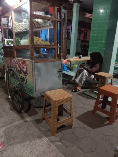 The image shows a food cart with a sign that says "Bakso" in Indonesian. The cart is parked on a sidewalk in front of a building with tiled walls. A person is sitting on a stool to the right of the cart, looking down at a mobile phone. The person is wearing a black hijab and a white shirt. There is a second, brown stool in front of the person. The cart is filled with various food items, such as noodles and meatballs. The image was taken in Kecamatan Brangsong, Indonesia, located at latitude: -6.943, longitude: 110.222.