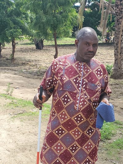 The image shows a man holding a white cane, suggesting he is visually impaired, outdoors in what appears to be a rural setting in Kabwir, Nigeria. He is wearing a long-sleeved patterned shirt in shades of maroon, gold, and blue, and holds a folded blue and white checkered cloth in his left hand. The man has a beard and his hair is graying. The background consists of dry, dusty ground with some sparse green vegetation and trees. In the distance, there are other people and what looks like cultivated fields. The lighting suggests it is daytime. The man's expression is neutral. The white cane is a mobility aid, indicating he is likely navigating the environment.