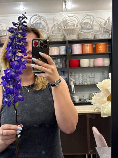 A woman is taking a selfie in a mirror, holding a stalk of purple delphinium flowers and showing off her light blue fingernails. She is wearing a dark gray, ribbed tank top and a diamond pendant necklace. Her blonde hair is shoulder-length. Behind her, shelves are filled with various items including wicker baskets, decorative round boxes in pink, orange, and red, and glass vases. The scene appears to be an indoor retail space, possibly a flower shop or a gift shop. The image does not provide specific geographical information, therefore its location in Assinovskaya, Russia, cannot be confirmed.