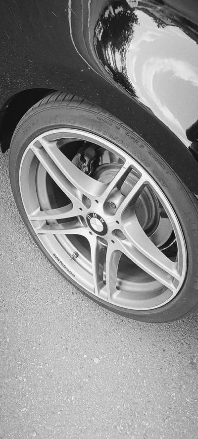 A close-up, black and white, medium shot captures the front passenger-side wheel of a black car, identified by the "BMW Performance" text on the rim. The wheel is a silver, multi-spoke alloy rim with the BMW logo prominently displayed in the center. The tire features various markings, including "OUTSIDE," "DOT UAD7," "TREADWEAR 300," "TRACTION AA," "TEMPERATURE A," and the tire size "245/35ZR18 92Y." The car's body, with its glossy, reflective surface, is visible in the upper portion of the image, showing streaks and some dirt. The wheel is positioned on a textured asphalt surface, common in urban environments. The overall scene is static and focuses on the automotive detail. The image is taken from a slightly elevated angle looking down at the wheel. No specific identifiable landmarks or context for Waiblingen, Germany are present within the image itself.