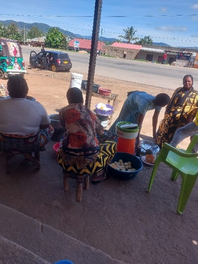 The scene depicts an outdoor roadside vending area in Kimamba, Tanzania, under a clear to partly cloudy sky. In the immediate foreground, a concrete surface transitions to dirt. Four adult females and a partial view of a fifth individual are present.
To the far left, a woman with her back to the camera is seated on a low wooden stool, wearing a white top and a patterned skirt. Central to the foreground, another woman, also with her back to the camera, sits on a stool adorned with yellow and black patterns, wearing a floral-patterned dress. Between these two seated figures, a dark pot is visible on the ground.
To the center-right, a woman in a light blue garment is bent over, attending to items on the ground, including an orange and white insulated container and a blue basin filled with numerous spherical food items, possibly fried dough. To her right, a green plastic chair is positioned. Further right, a woman in a black and yellow patterned dress faces forward, smiling. Part of another person's leg and clothing are visible next to her.
In the mid-ground, a dirt shoulder parallels an asphalt road. A small wooden table with various containers, including white and red bowls and a white plastic bucket, is situated behind the bent-over woman. On the dirt shoulder, a dark-colored SUV with all doors open is parked, featuring distinct yellow wheel rims. To the far left mid-ground, a green and white three-wheeled vehicle, identified as a Bajaji or Tuk-Tuk, decorated with religious images, is parked.
The background features additional buildings along the roadside, including structures with red corrugated roofs and a partially constructed or dilapidated brick building. Utility poles with wires are visible. Rolling hills or mountains rise in the distant background.