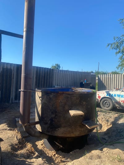 The image depicts an outdoor scene on a sunny day, under a clear blue sky. In the foreground, a large, dark, metal vat is partially buried in the ground, with a metal pipe extending from its side and upward. There are two small wooden platforms attached to the vat. A green hose snakes around the base of the vat.
Behind the vat, a corrugated metal fence runs horizontally across the frame. To the right of the vat, a white car is parked. The car has Cyrillic text painted on its side, including the word "ХУДОЖЕСТВЕННАЯ," which translates to "artistic" or "art."
The ground appears to be sandy and dry, with some sparse vegetation. The overall impression is of a rustic, possibly industrial or workshop setting in a rural or suburban area. The location is identified as Mamontovo, Russia. No people are visible in the image.
