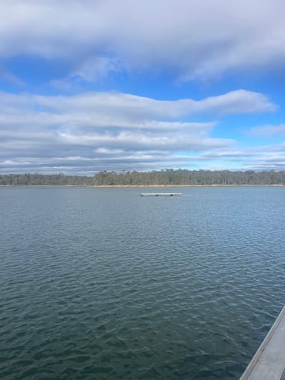 The image depicts a wide, calm body of water, likely a lake or reservoir, under a cloudy blue sky. The water's surface is slightly rippled, reflecting the light and creating a textured appearance. In the middle distance, a rectangular floating platform is visible on the water. The horizon is lined with a dense forest of eucalyptus trees, characteristic of the Australian landscape, suggesting this location could be in or near Kangaroo Flat, Australia, a region known for its natural bushland and waterways. The foreground shows the edge of a dock or pier, made of grey material. The overall atmosphere is serene and natural, with the vastness of the water and sky dominating the scene.