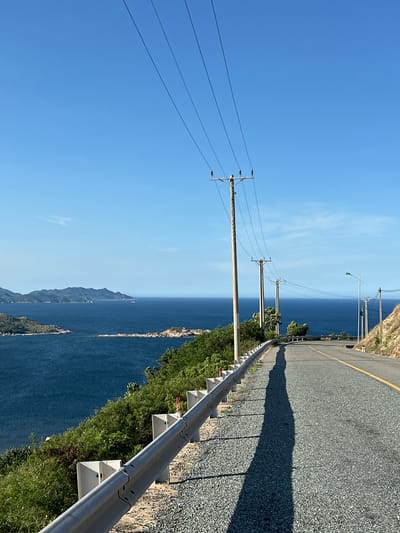 This is a photograph taken on a sunny day of a coastal road in the city of Vĩnh Hải, Vietnam. The road curves to the right, leading the viewer's eye towards the horizon. On the left side of the road, a metal guardrail separates the asphalt from a steep, vegetated embankment. The embankment is covered in lush green bushes and small trees, with some rocky outcrops visible. Beyond the embankment, a vast expanse of deep blue ocean stretches out, dotted with several small, green islands in the distance. The sky above is a clear, bright blue with a few wispy white clouds. A series of utility poles with power lines run along the right side of the road, casting long shadows on the asphalt due to the bright sunlight. A streetlamp can be seen further down the road. The road surface is asphalt, with a yellow line marking the center. The scene depicts a scenic and tranquil coastal drive.