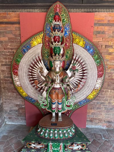 The image depicts a large, intricate sculpture of a deity, likely a Buddhist or Hindu figure, mounted on a wall in a building in the city of Lalitpur, Nepal. The sculpture is multi-armed, with a detailed and colorful head, torso, and legs. The hands radiate outwards from the body in a fan-like arrangement. The background of the sculpture is a semi-circular design with a textured, silver-colored interior and a decorative border with vibrant colors such as red, green, yellow, and blue. The wall behind the sculpture is constructed of red bricks, and there is a red panel behind part of the circular design. To the right of the figure is part of a dark green base platform embellished with similar colorful patterns. The overall setting suggests an interior display within a temple, museum, or another cultural location in Lalitpur.