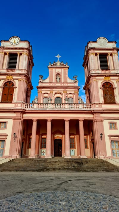 The image depicts the facade of Göttweig Abbey, a Benedictine monastery located in Furth bei Göttweig, Austria. The abbey is characterized by its Baroque architecture, with a central pink and cream-colored structure flanked by two towers. Each tower features a clock face and ornate detailing. A large set of stone stairs leads up to the main entrance, which is framed by colossal columns. The sky above is a clear, vibrant blue. There are no people visible in the image. The foreground shows a cobbled circular area and asphalt, suggesting a public square or entrance.