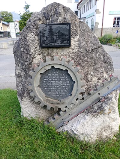 The image shows a large rock with a metal gear and a plaque attached to it. The gear is large and appears to be made of metal with red accents on its teeth. Inside the gear, a dark plaque contains text and an image. The plaque above the gear features a black and white engraved image of a building with a mountain and trees in the background. The rock is situated on a grassy area with pavement and buildings visible in the background. The scene is outdoors, suggesting a public space. The plaque's inscription, if legible, would provide specific details about the monument's purpose or historical significance. Based on available information, this monument is located in Brannenburg, Germany, and commemorates the Wendelsteinbahn cog railway, which operated from 1912 to 1961.