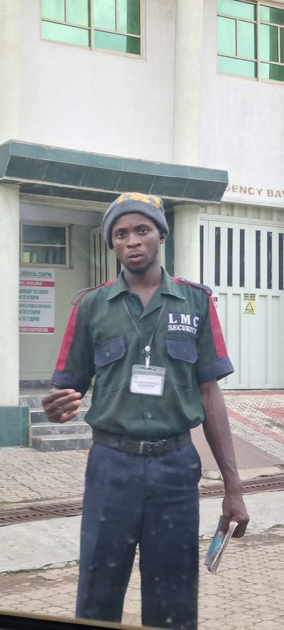 A man wearing a dark green security uniform with red and black accents on the sleeves and collar stands outside a building. He has a gray beanie on his head and a lanyard with an ID badge around his neck. The badge is visible and appears to have text and a photo on it. The man is holding a folded pamphlet or booklet in his right hand. The building behind him is white with green-tinted windows and a sign that reads "EMERGENCY BAY." Another sign on the building lists "LMC MEDICAL CENTRE OPERATING HOURS." The ground in front of the building is paved with interlocking stones and has some metal grates. The scene appears to be in a public area.