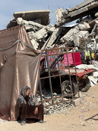 The image depicts a scene of extensive urban destruction in Jabalia, Palestinian Territory, under a clear daytime sky. In the foreground, an elderly individual is seated directly on the barren dirt ground, leaning against a makeshift shelter constructed from a shiny, coppery-brown tarp material secured with straps. This individual wears a multi-patterned head covering and a long, dark brown outer garment over a patterned shirt; their right hand is raised to their forehead, partially obscuring their face, while their left hand rests on their lap.
To the immediate right of the seated person, a rusty, rectangular metal frame, possibly from a bed or window grill, stands upright. Behind this frame, a red-framed utility cart with two visible wheels is positioned on uneven ground littered with small concrete debris. The cart is laden with various items, including patterned blankets and a red towel draped over its structure, and several articles of clothing (e.g., t-shirts, bags) hanging on a line or structure behind it. White tarpaulins are visible behind the cart.
The background is dominated by a massive pile of shattered concrete slabs and twisted rebar, indicating the remains of a demolished building. Amidst this rubble, a second individual, wearing a greenish top, is partially visible near the upper portion of the debris pile, appearing to be observing or navigating the destruction.